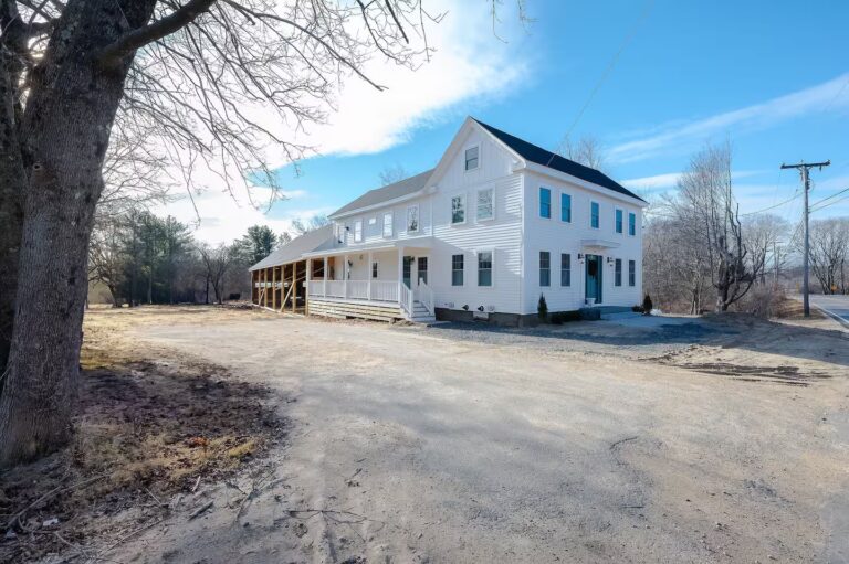 Photo: exterior shot of a white farmhouse with muddy ground and bare trees in the foreground.